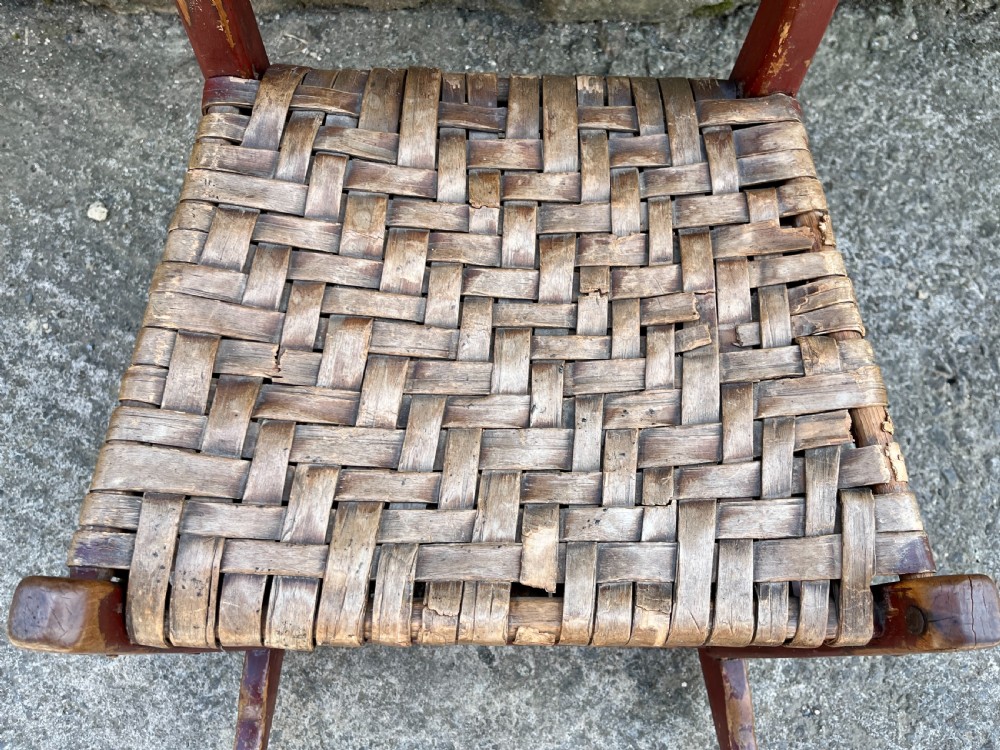 A primitive red-painted wooden rocking chair featuring a woven splint seat and scalloped back slats. View XII