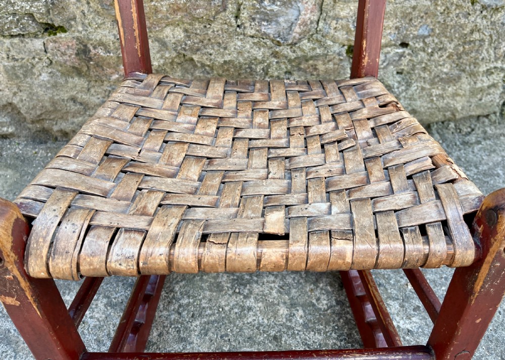 A primitive red-painted wooden rocking chair featuring a woven splint seat and scalloped back slats. View XIII