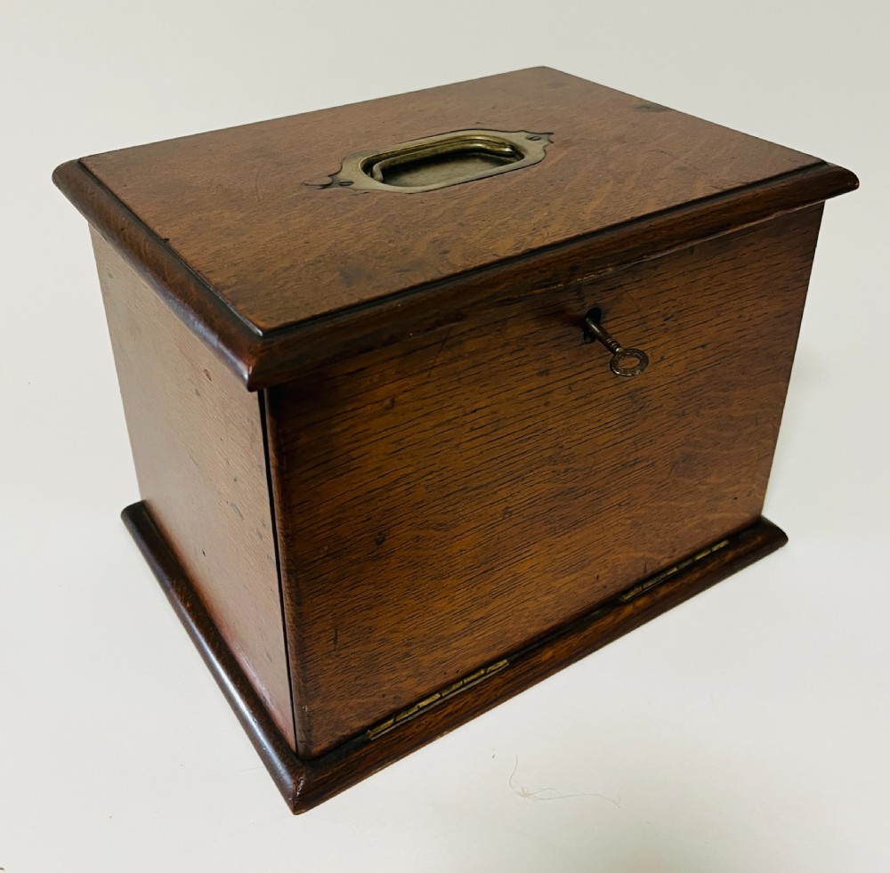 An antique oak stationery cabinet featuring a flush brass top handle, a brass keyhole with a key, and a hinged front panel. View XI