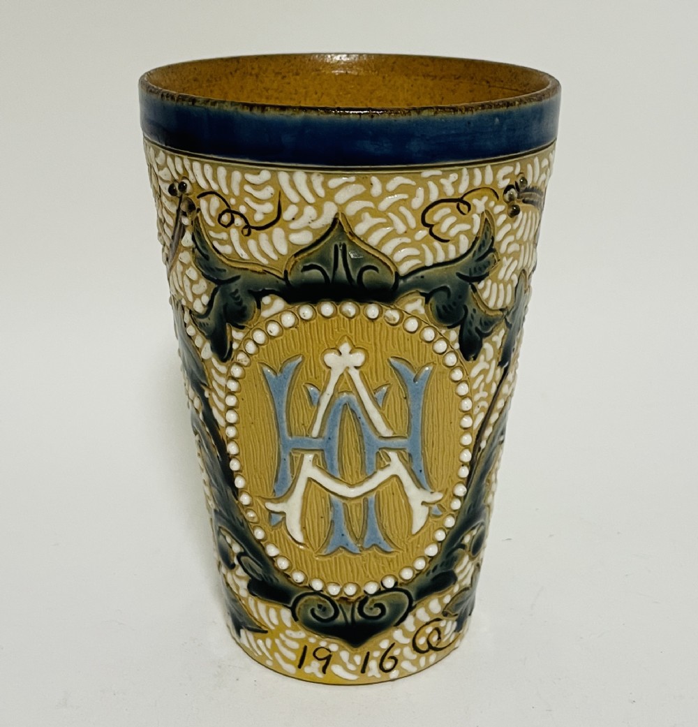 An Arts and Crafts salt-glazed stoneware beaker from 1916 featuring a central monogram framed by white beaded slip, blue detailing, and an ochre textured background.