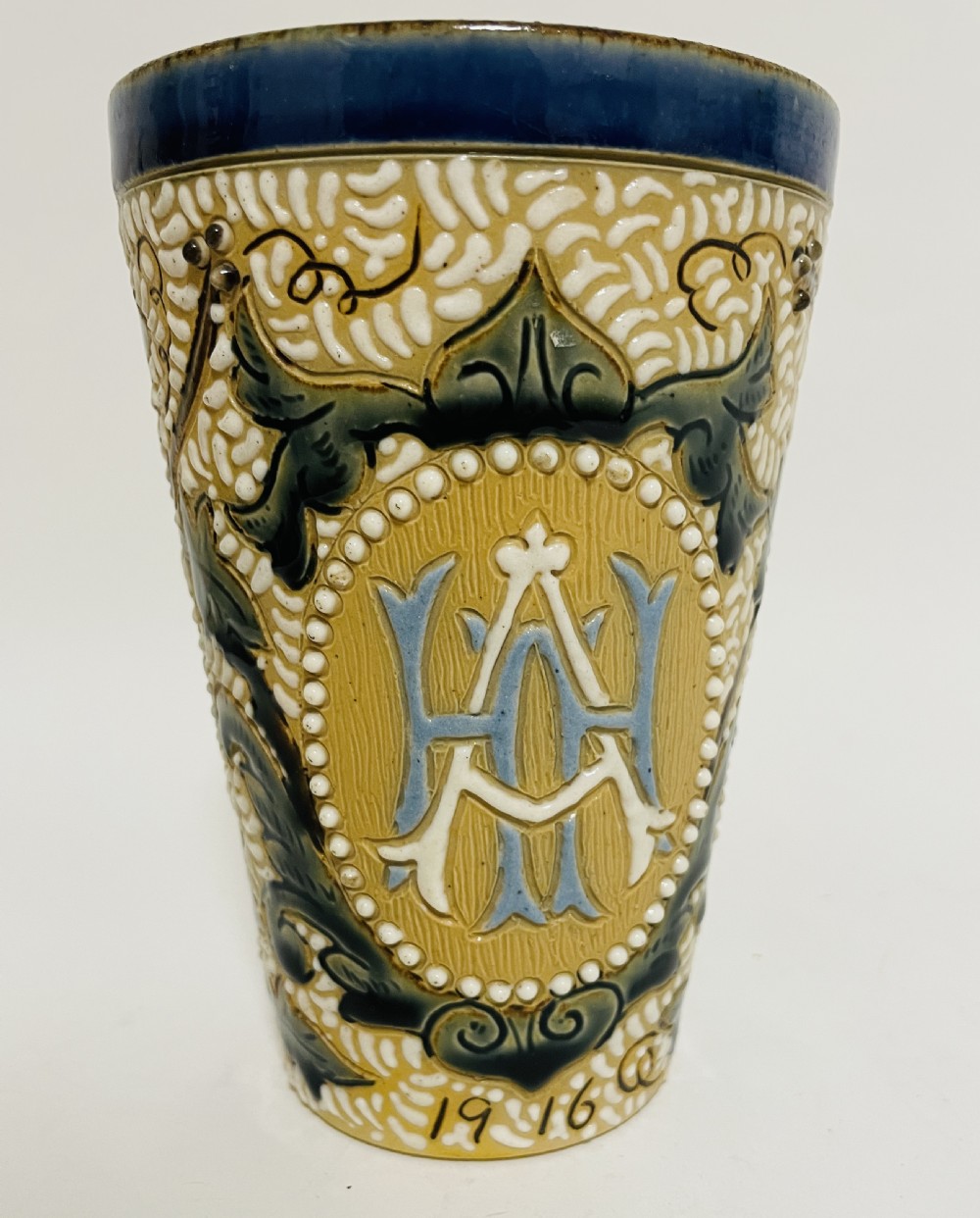 An Arts and Crafts salt-glazed stoneware beaker from 1916 featuring a central monogram framed by white beaded slip, blue detailing, and an ochre textured background. View VII