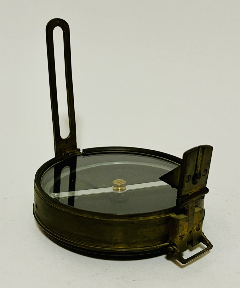 An antique brass surveying compass featuring folding sight vanes, a central needle, and a glass-covered dial.