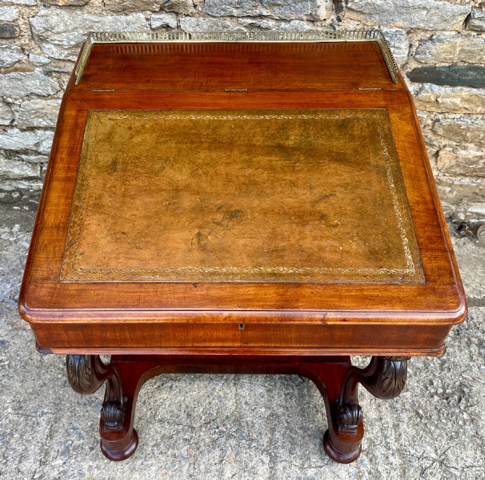 A Victorian mahogany Davenport desk featuring a sloped leather writing surface, side drawers with turned wooden knobs, a top brass gallery rail, and carved scroll front supports. View II