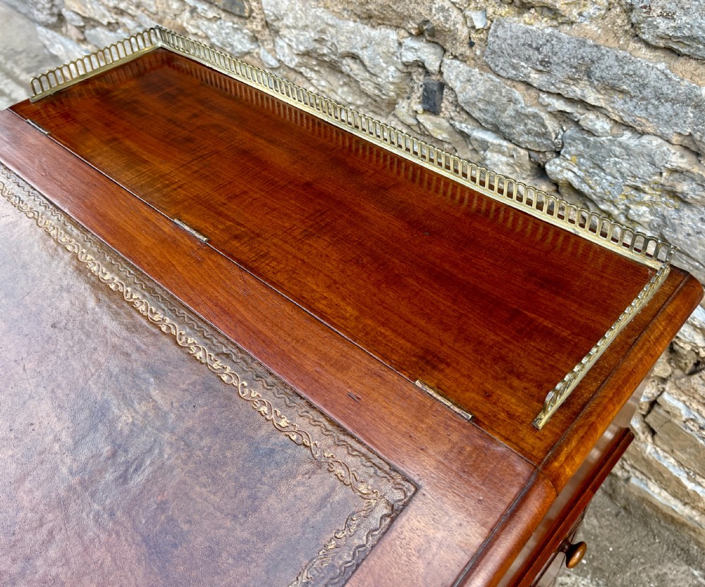 A Victorian mahogany Davenport desk featuring a sloped leather writing surface, side drawers with turned wooden knobs, a top brass gallery rail, and carved scroll front supports. View IV