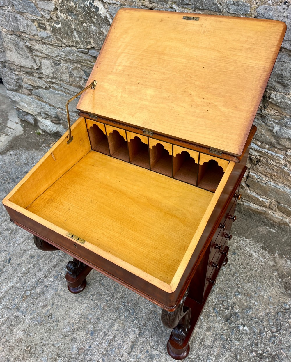 A Victorian mahogany Davenport desk featuring a sloped leather writing surface, side drawers with turned wooden knobs, a top brass gallery rail, and carved scroll front supports. View IX