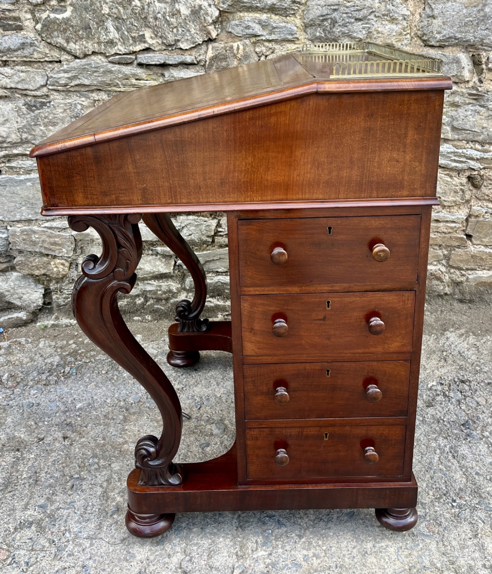 A Victorian mahogany Davenport desk featuring a sloped leather writing surface, side drawers with turned wooden knobs, a top brass gallery rail, and carved scroll front supports. View X