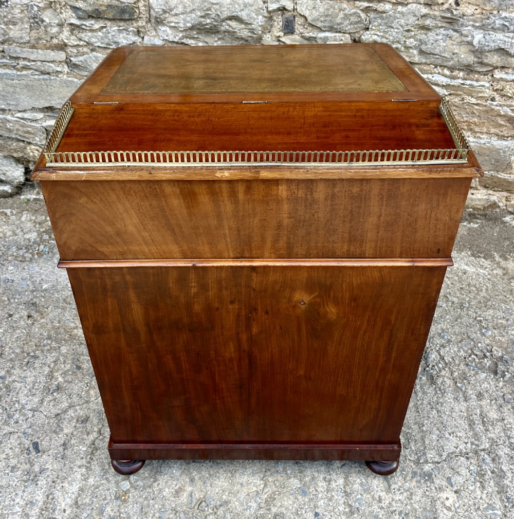A Victorian mahogany Davenport desk featuring a sloped leather writing surface, side drawers with turned wooden knobs, a top brass gallery rail, and carved scroll front supports. View XII