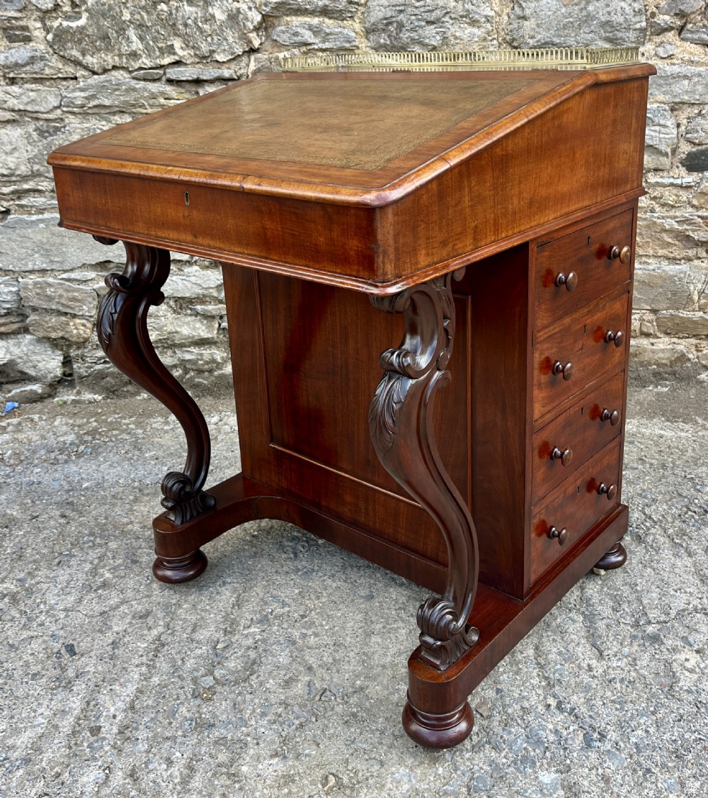 A Victorian mahogany Davenport desk featuring a sloped leather writing surface, side drawers with turned wooden knobs, a top brass gallery rail, and carved scroll front supports. View XIII