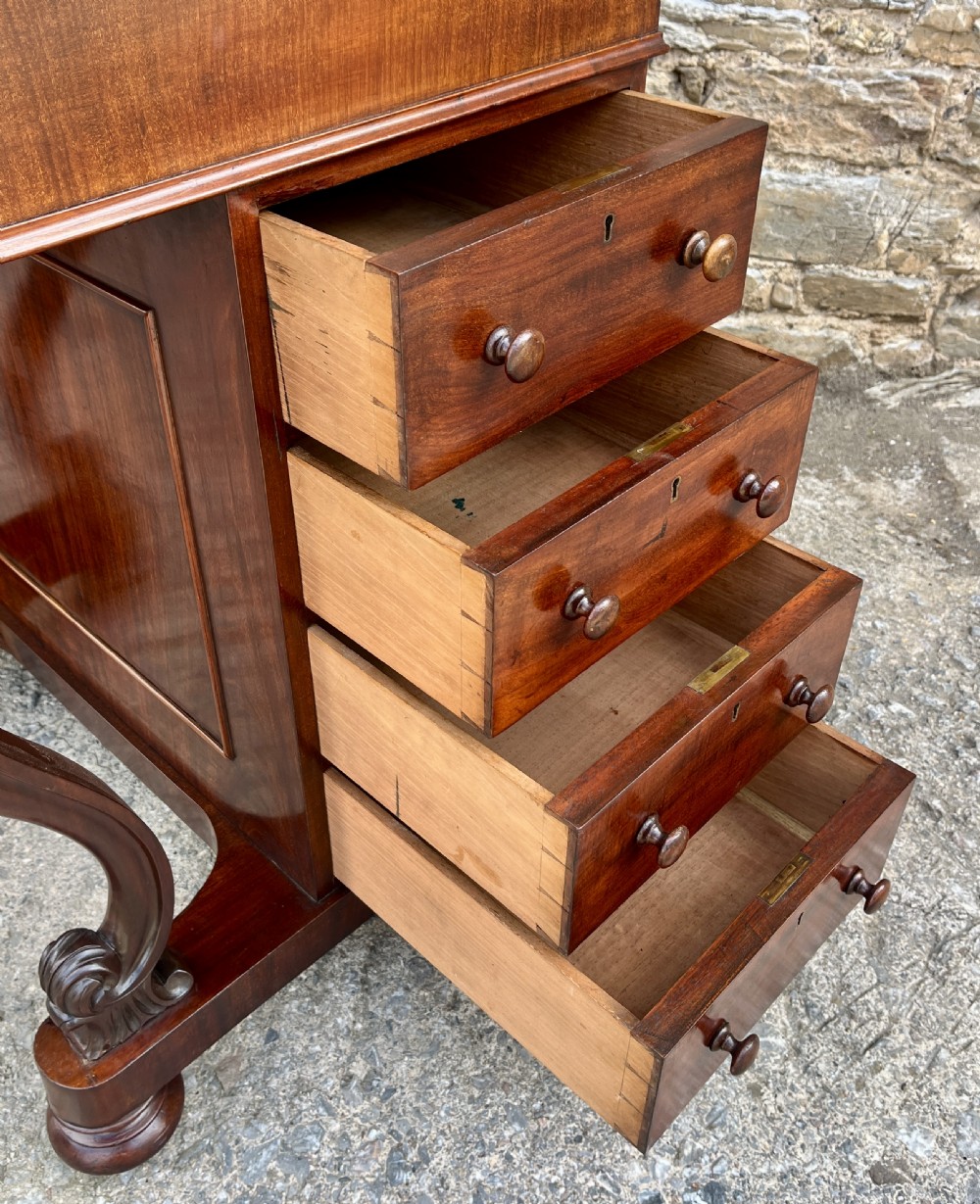 A Victorian mahogany Davenport desk featuring a sloped leather writing surface, side drawers with turned wooden knobs, a top brass gallery rail, and carved scroll front supports. View XIV