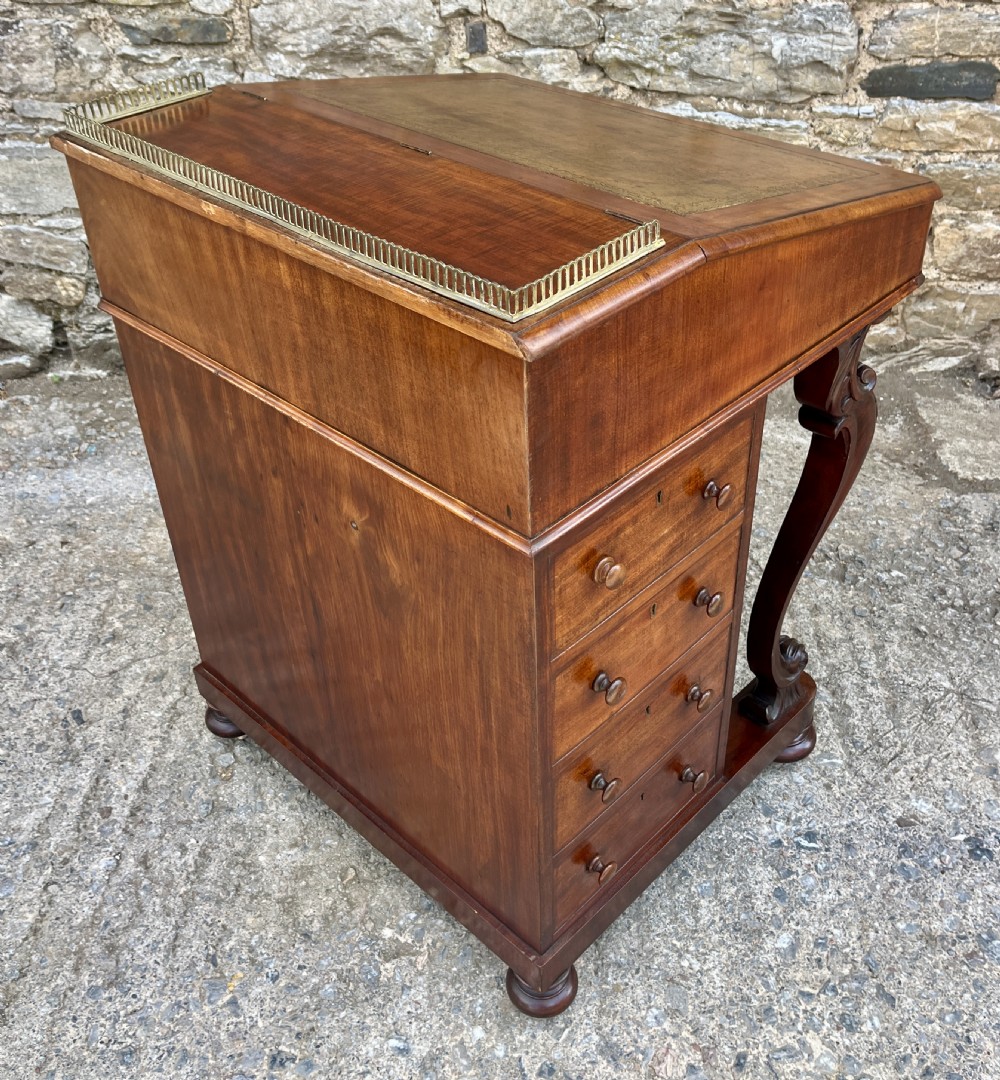 A Victorian mahogany Davenport desk featuring a sloped leather writing surface, side drawers with turned wooden knobs, a top brass gallery rail, and carved scroll front supports. View XVI