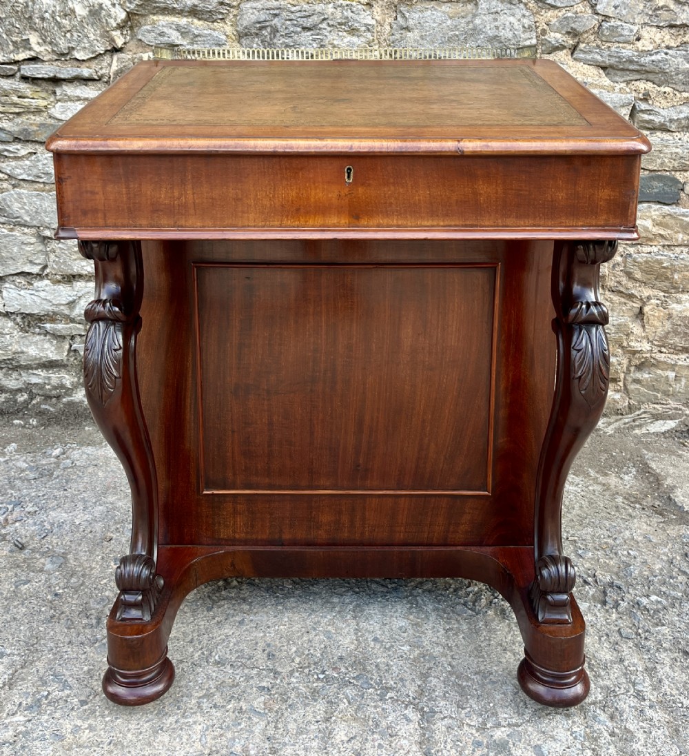 A Victorian mahogany Davenport desk featuring a sloped leather writing surface, side drawers with turned wooden knobs, a top brass gallery rail, and carved scroll front supports. View XVII