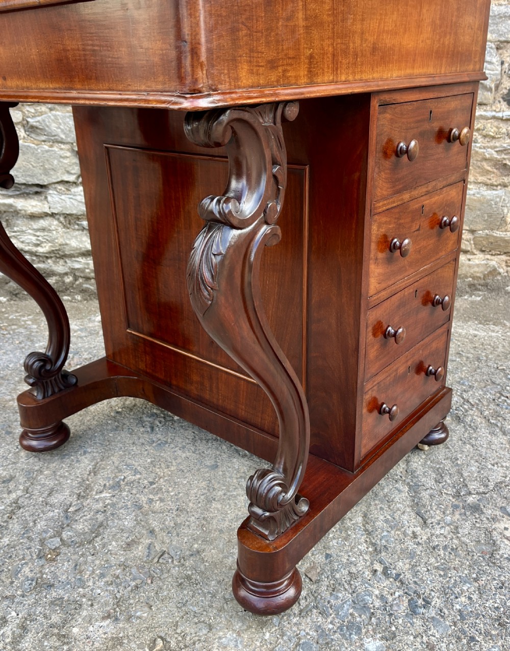 A Victorian mahogany Davenport desk featuring a sloped leather writing surface, side drawers with turned wooden knobs, a top brass gallery rail, and carved scroll front supports. View XVIII