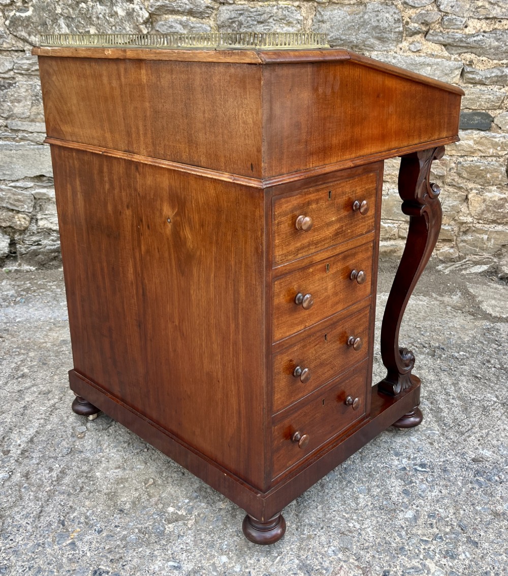 A Victorian mahogany Davenport desk featuring a sloped leather writing surface, side drawers with turned wooden knobs, a top brass gallery rail, and carved scroll front supports. View XIX