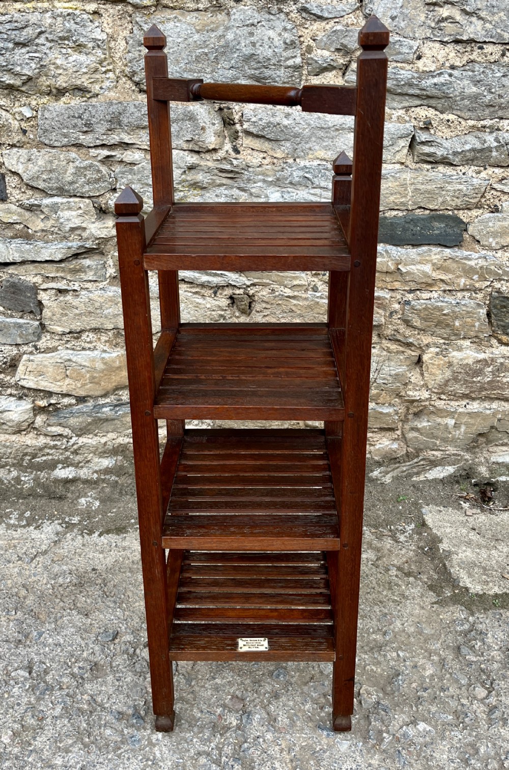 A tall, four-tier dark teak ships whatnot stand featuring slatted timber shelves, pyramid finials, a top carrying rail, and a brass makers plaque on the lowest shelf.