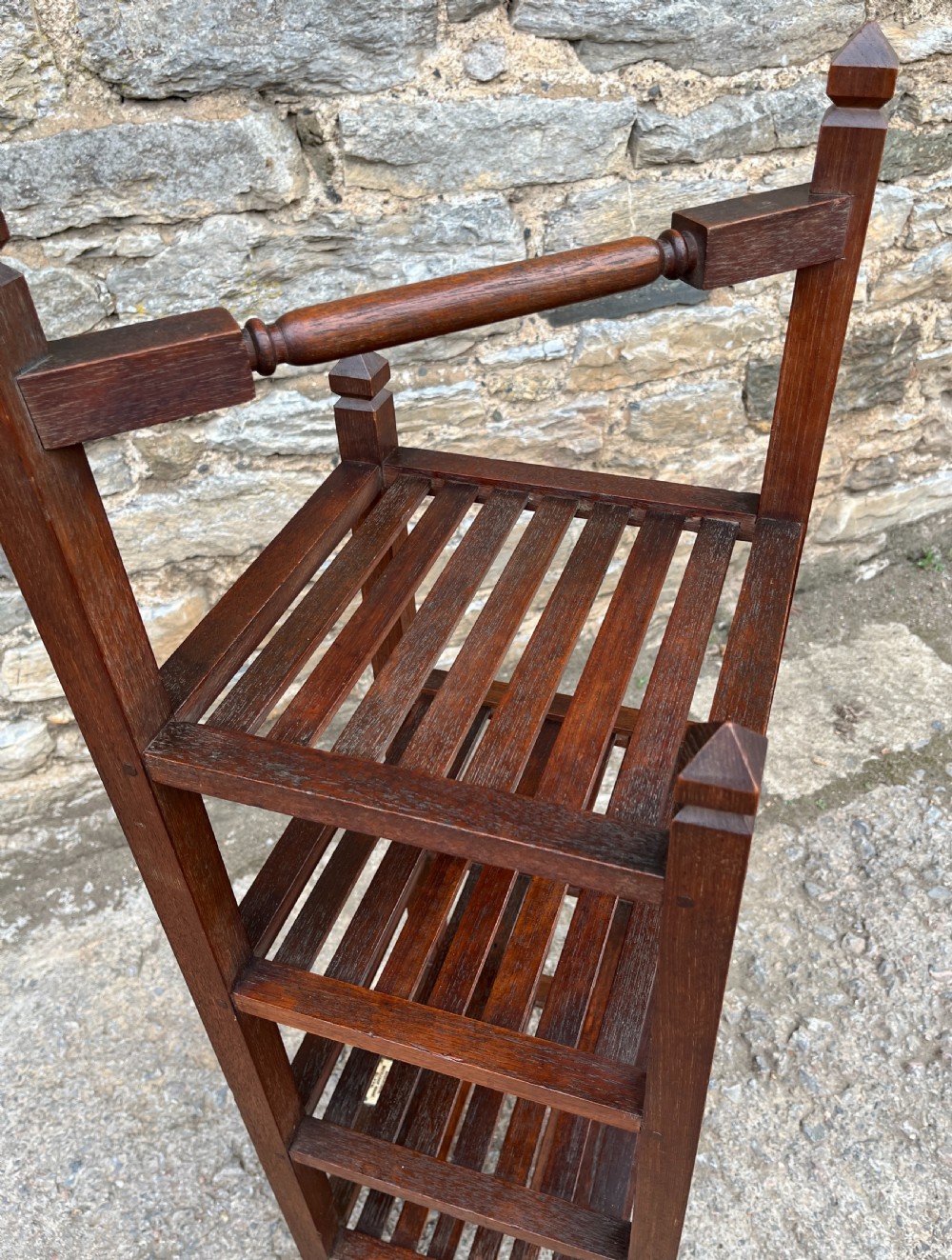 A tall, four-tier dark teak ships whatnot stand featuring slatted timber shelves, pyramid finials, a top carrying rail, and a brass makers plaque on the lowest shelf. View X
