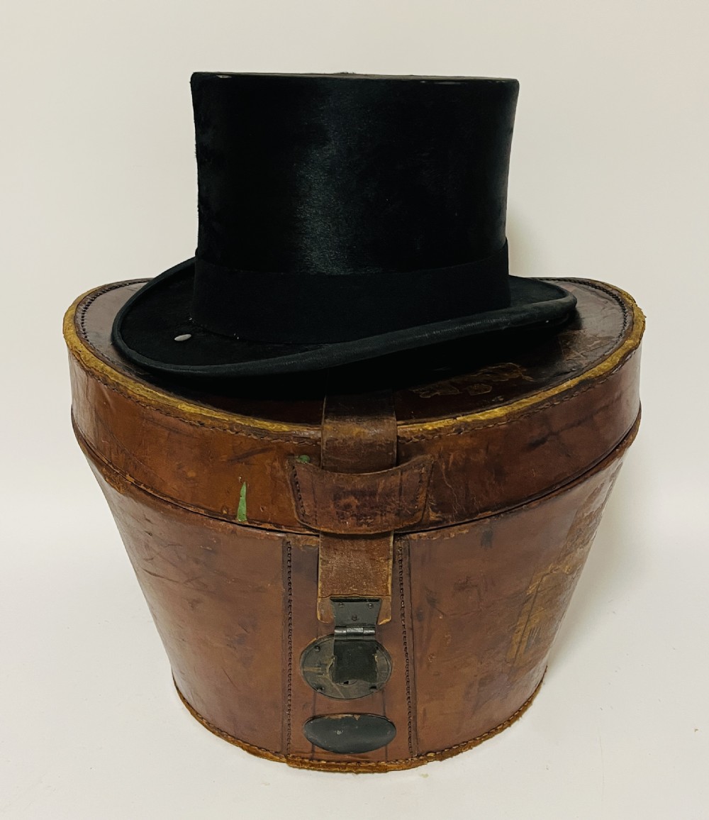 A black silk plush top hat resting on top of a bucket-shaped tan leather carrying case with a metal lock.