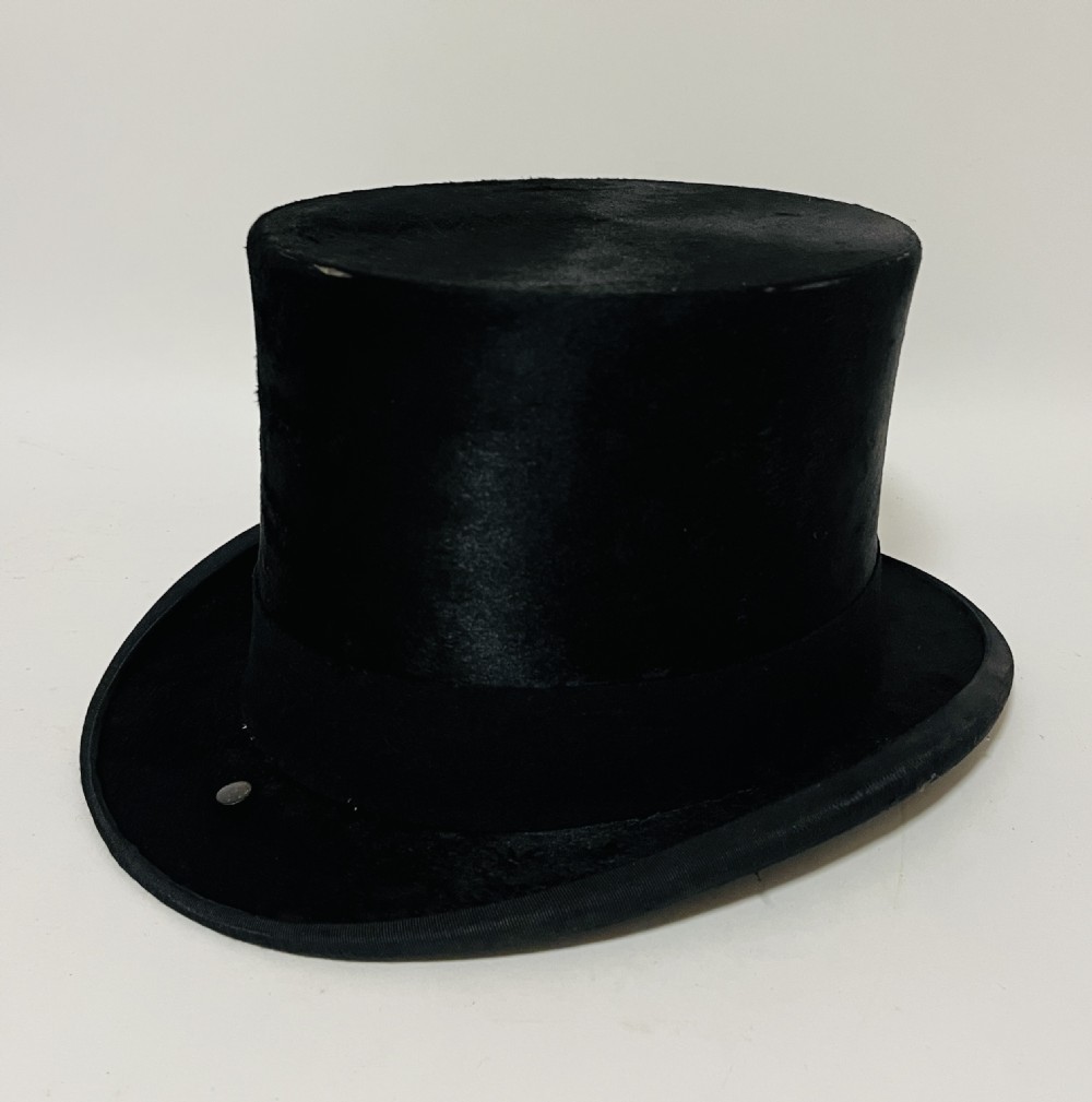A black silk plush top hat resting on top of a bucket-shaped tan leather carrying case with a metal lock. View II
