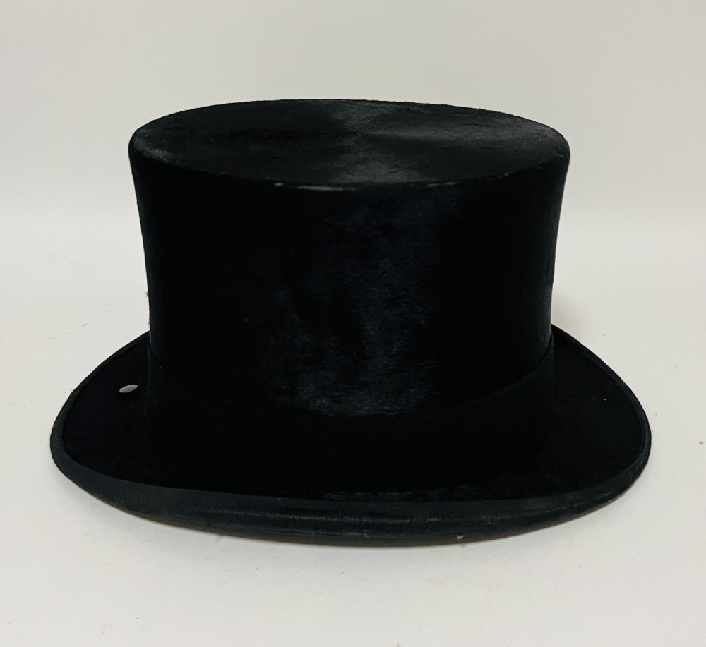 A black silk plush top hat resting on top of a bucket-shaped tan leather carrying case with a metal lock. View III