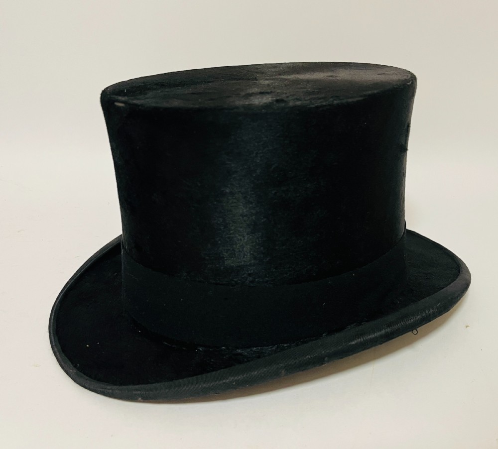 A black silk plush top hat resting on top of a bucket-shaped tan leather carrying case with a metal lock. View V