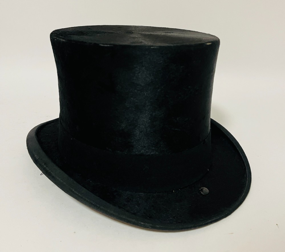 A black silk plush top hat resting on top of a bucket-shaped tan leather carrying case with a metal lock. View VI