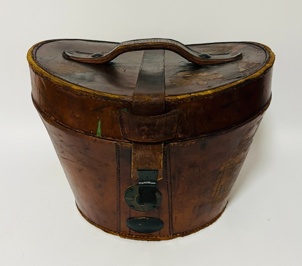 A black silk plush top hat resting on top of a bucket-shaped tan leather carrying case with a metal lock. View IX