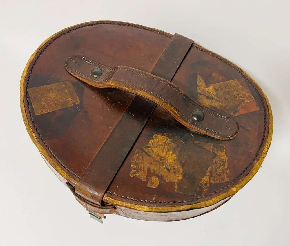 A black silk plush top hat resting on top of a bucket-shaped tan leather carrying case with a metal lock. View X