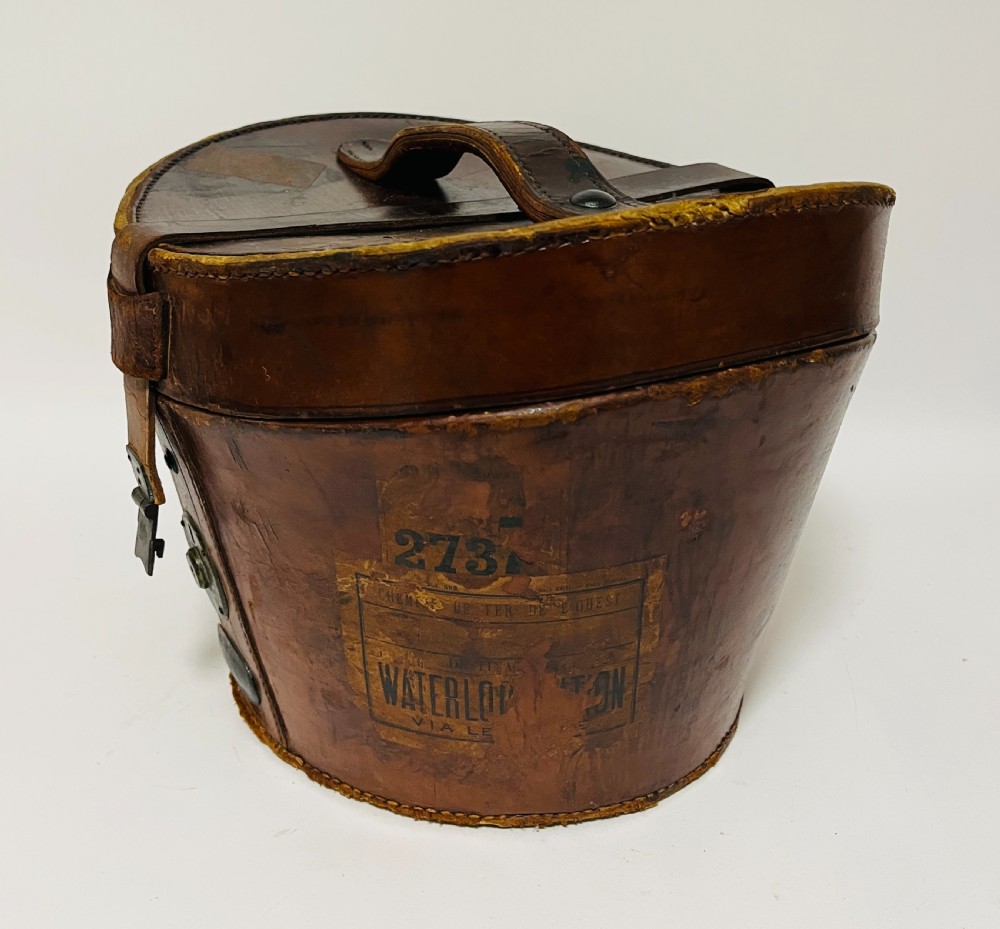 A black silk plush top hat resting on top of a bucket-shaped tan leather carrying case with a metal lock. View XI