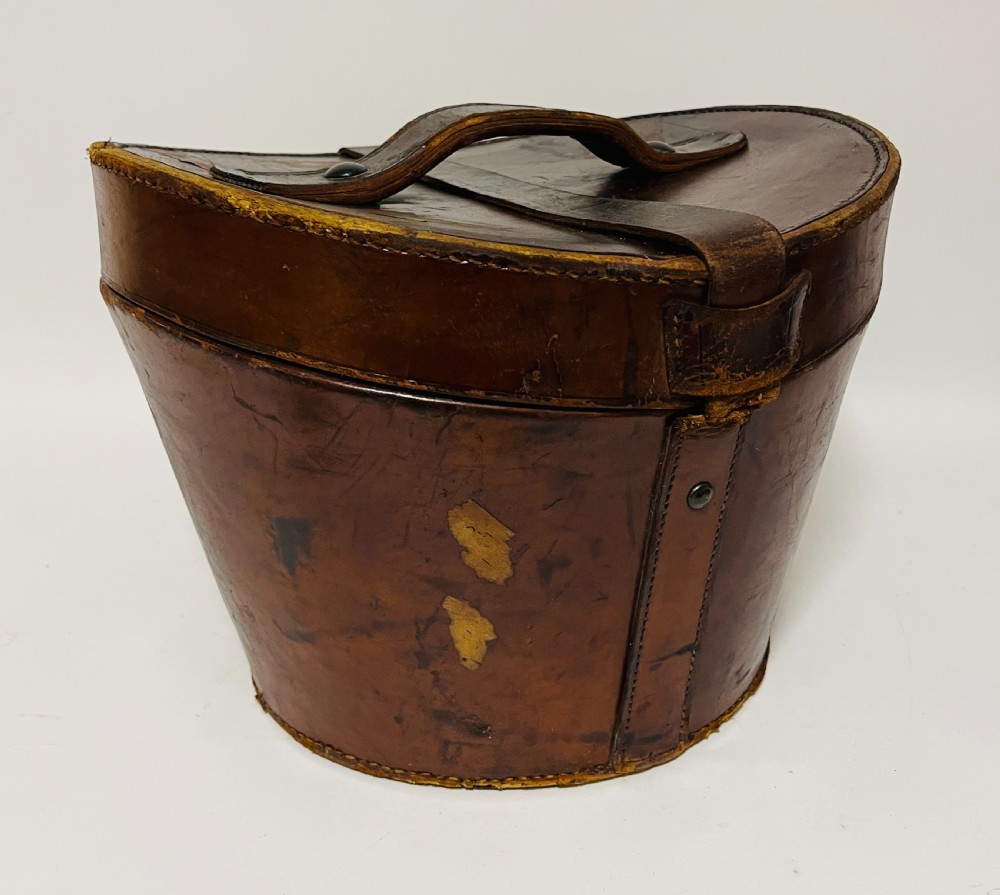 A black silk plush top hat resting on top of a bucket-shaped tan leather carrying case with a metal lock. View XII