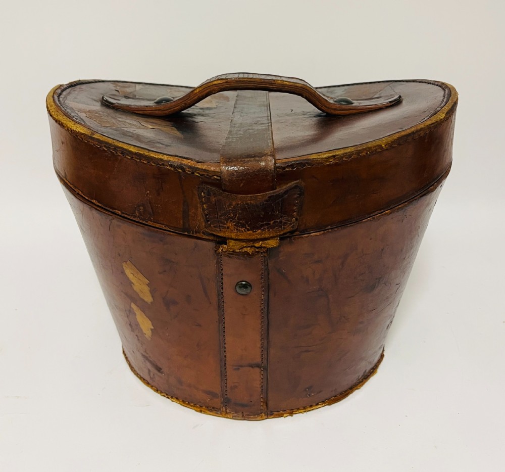 A black silk plush top hat resting on top of a bucket-shaped tan leather carrying case with a metal lock. View XIII