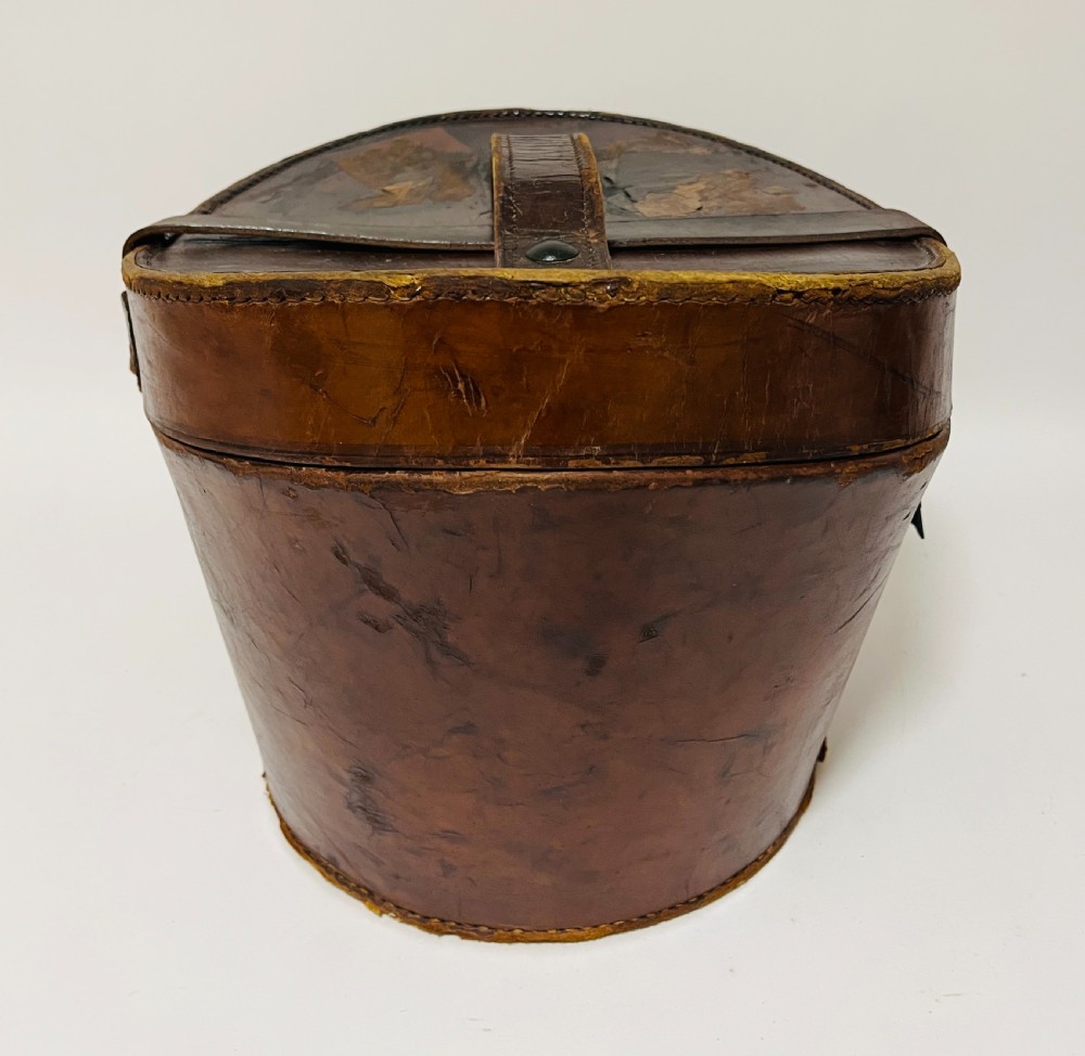 A black silk plush top hat resting on top of a bucket-shaped tan leather carrying case with a metal lock. View XIV