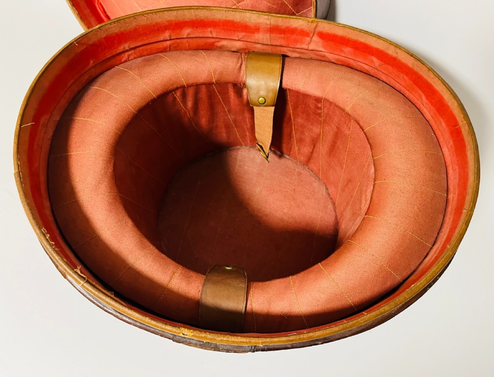 A black silk plush top hat resting on top of a bucket-shaped tan leather carrying case with a metal lock. View XV