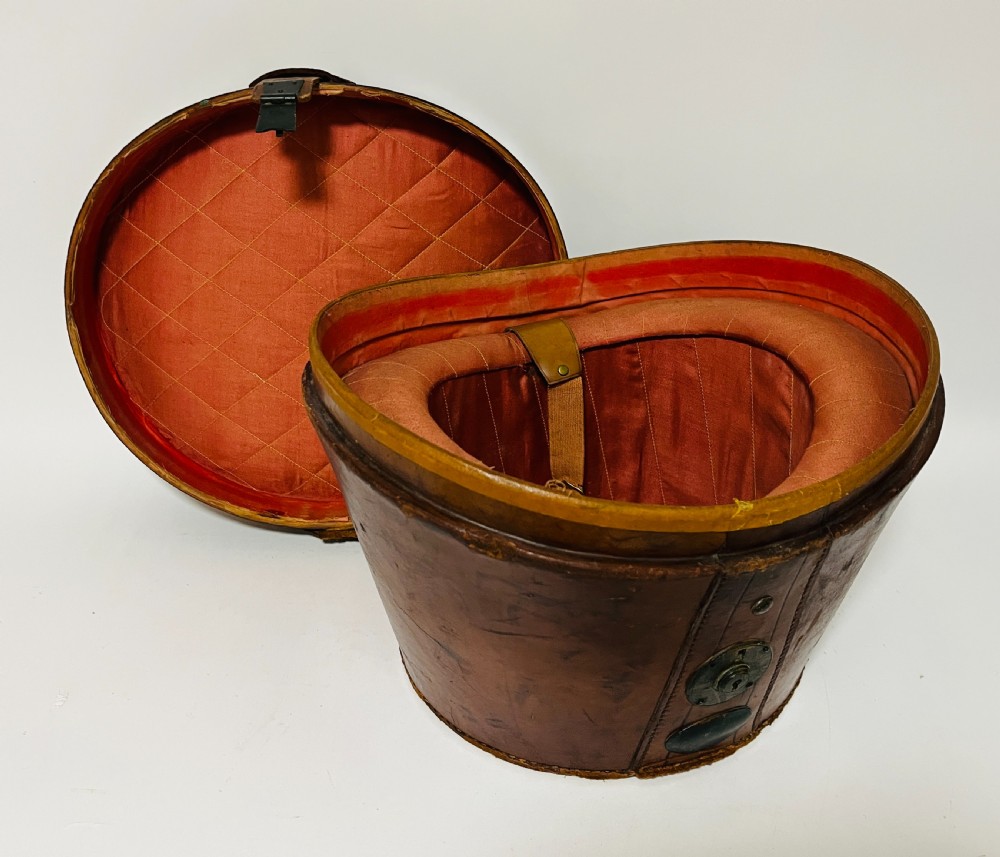 A black silk plush top hat resting on top of a bucket-shaped tan leather carrying case with a metal lock. View XVI