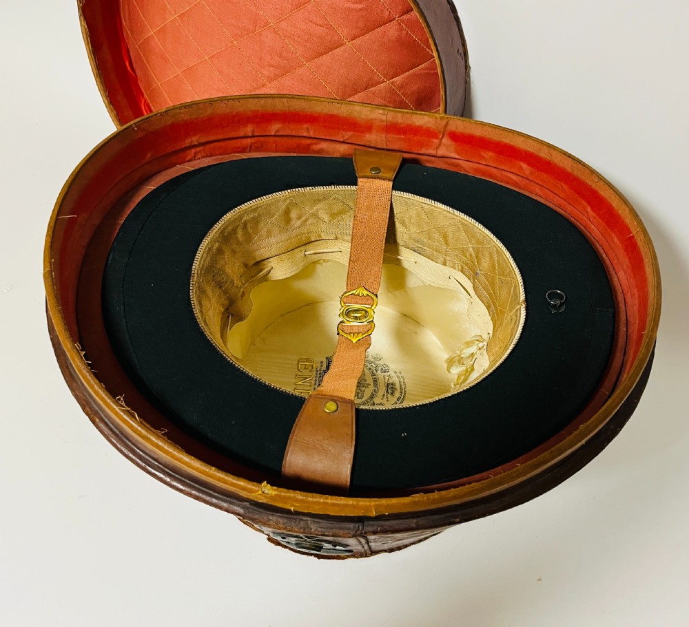 A black silk plush top hat resting on top of a bucket-shaped tan leather carrying case with a metal lock. View XVII