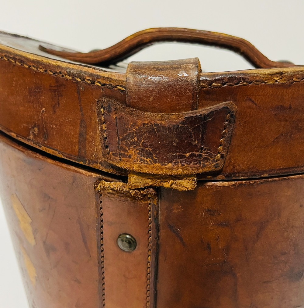 A black silk plush top hat resting on top of a bucket-shaped tan leather carrying case with a metal lock. View XVIII