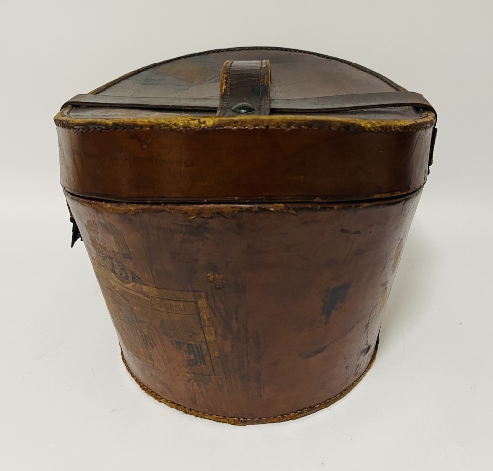 A black silk plush top hat resting on top of a bucket-shaped tan leather carrying case with a metal lock. View XIX