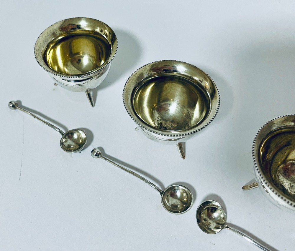 A set of four circular silver cauldron-shaped open salt cellars with beaded rims, tripod feet, and matching miniature ball-top spoons. View V