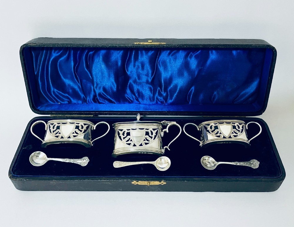 A three-piece Victorian silver condiment set with matching spoons displayed inside a fitted blue satin-lined dark presentation case.