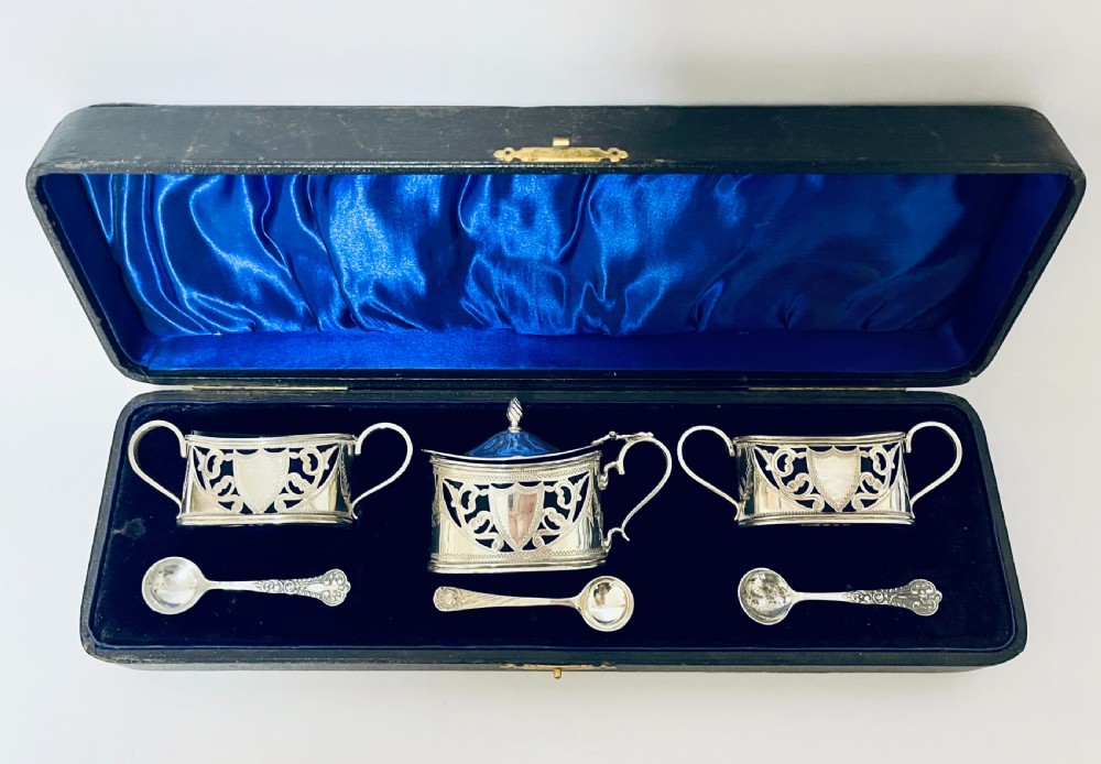 A three-piece Victorian silver condiment set with matching spoons displayed inside a fitted blue satin-lined dark presentation case. View II