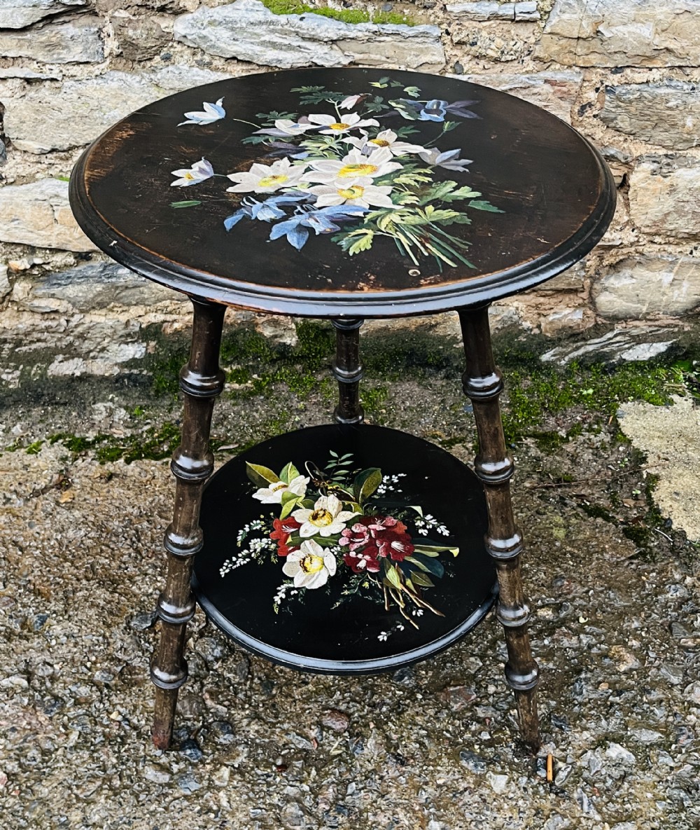 A circular two-tiered ebonised wooden side table featuring turned bobbin legs and hand-painted floral arrangements on both levels.