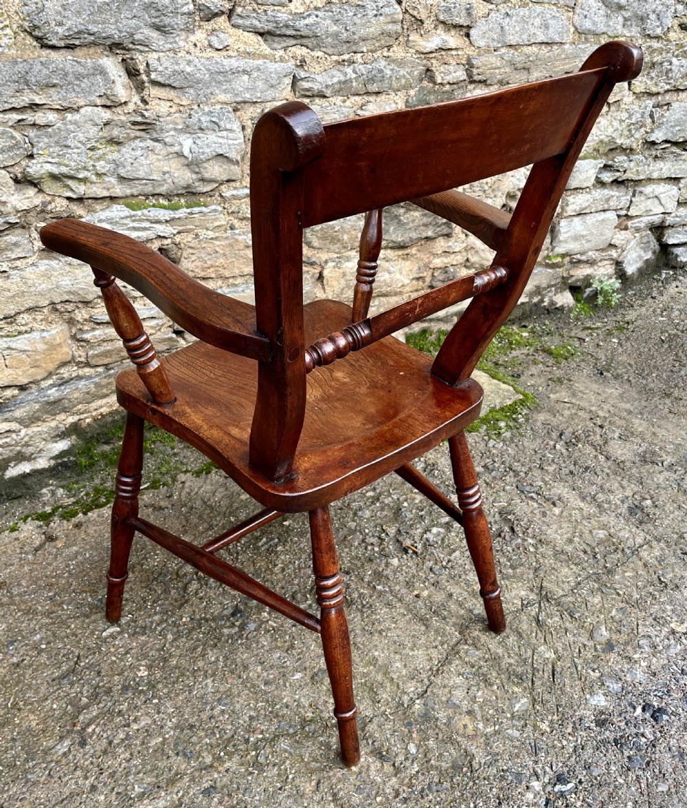 An antique wooden bar-back carver chair featuring curved armrests, turned spindle supports, a shaped seat with visible wood grain, and a warm brown finish. View VI