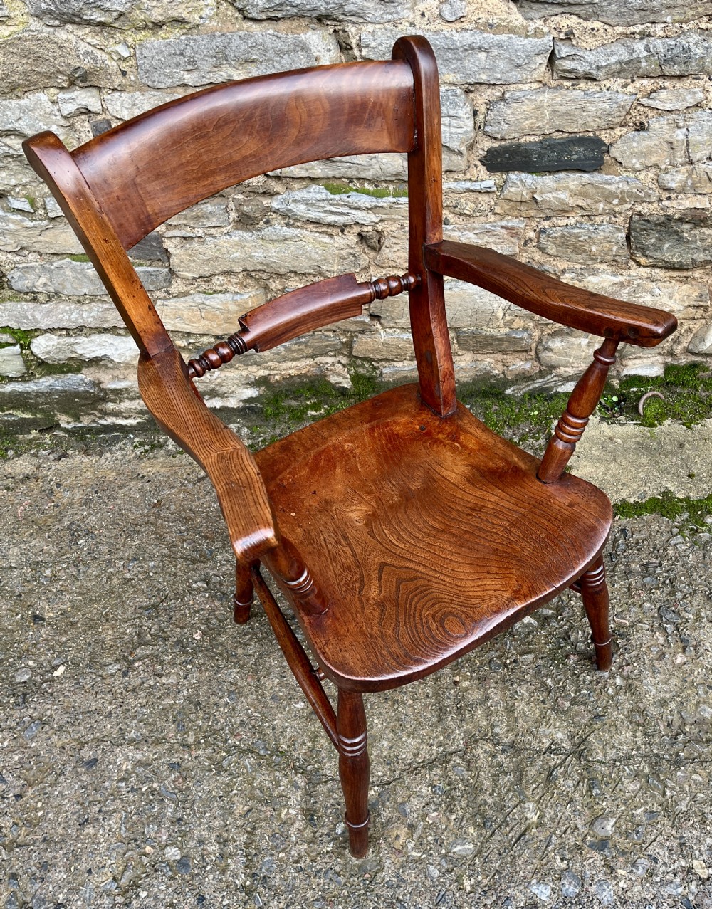 An antique wooden bar-back carver chair featuring curved armrests, turned spindle supports, a shaped seat with visible wood grain, and a warm brown finish. View X