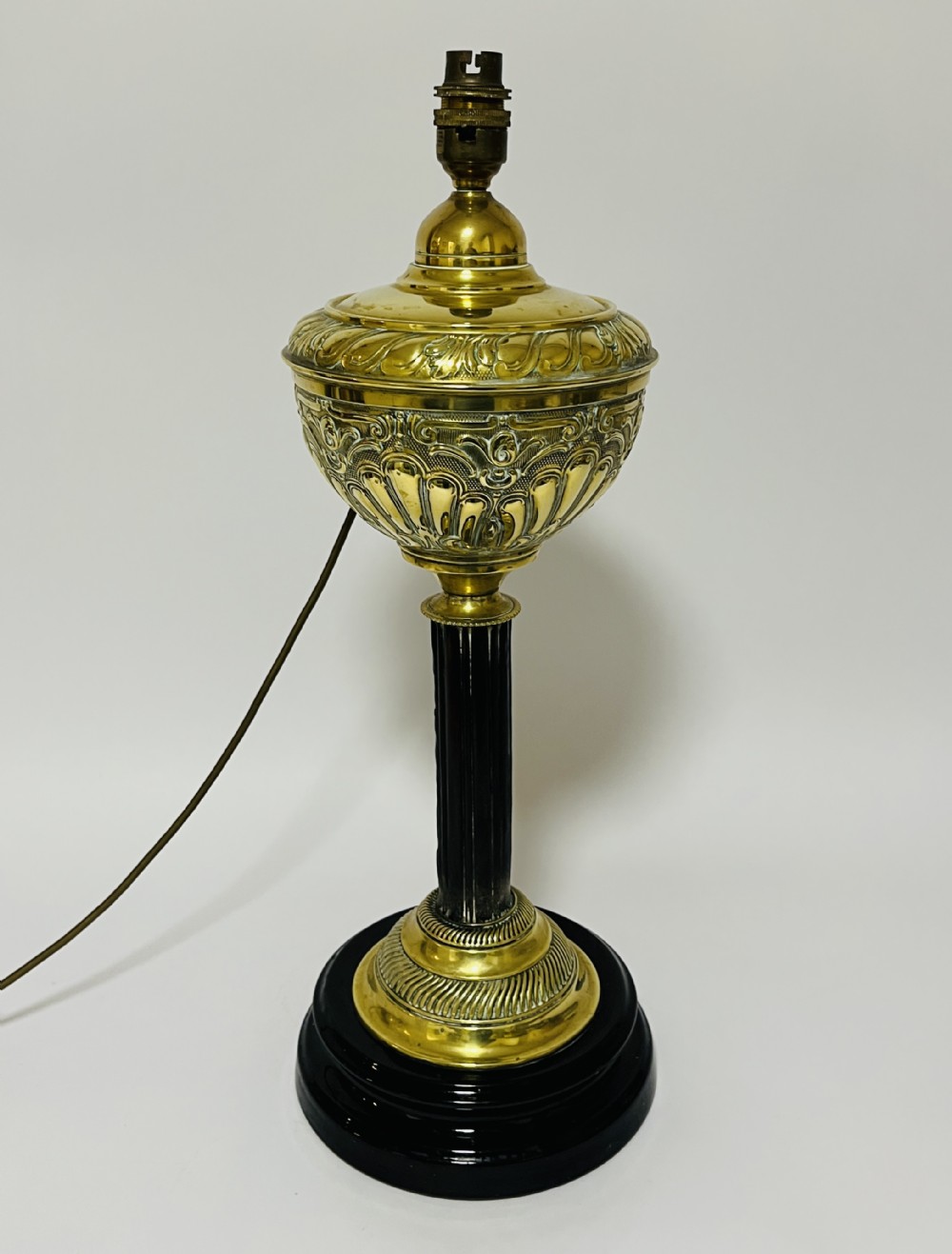 An antique Victorian embossed brass oil lamp converted into an electric table lamp with a reeded column and black circular base.