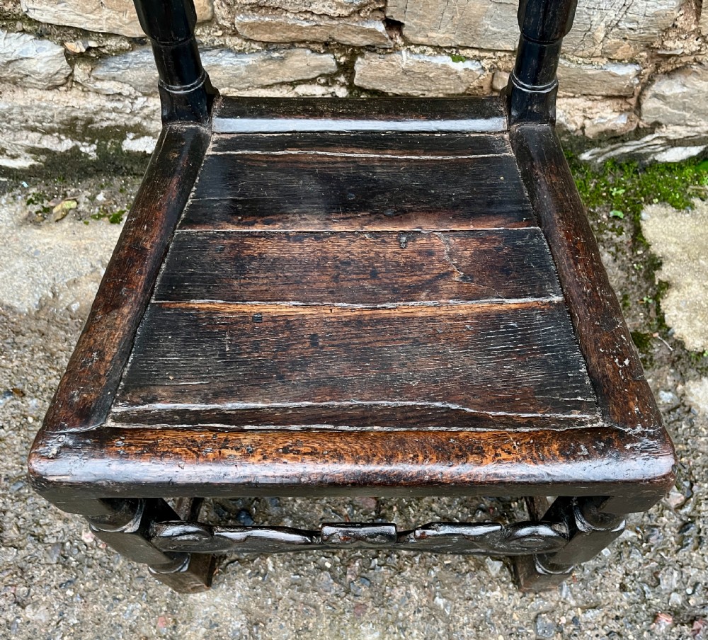 A dark 17th-century carved oak backstool with barley-twist uprights, a carved cresting rail, vertical back splats, a solid seat, and turned legs. View II