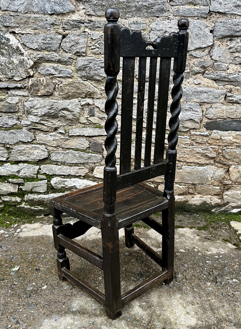 A dark 17th-century carved oak backstool with barley-twist uprights, a carved cresting rail, vertical back splats, a solid seat, and turned legs. View V