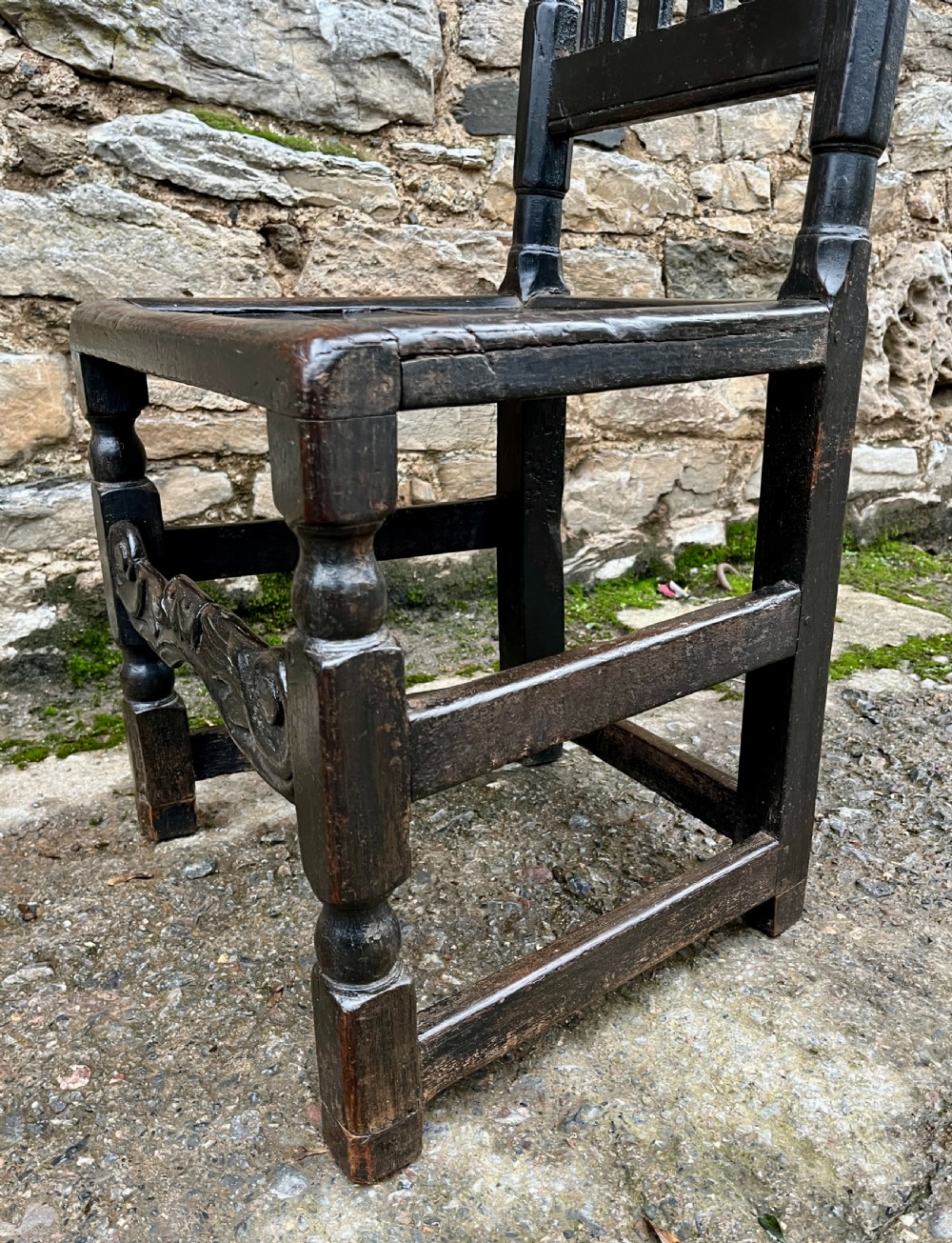 A dark 17th-century carved oak backstool with barley-twist uprights, a carved cresting rail, vertical back splats, a solid seat, and turned legs. View VI