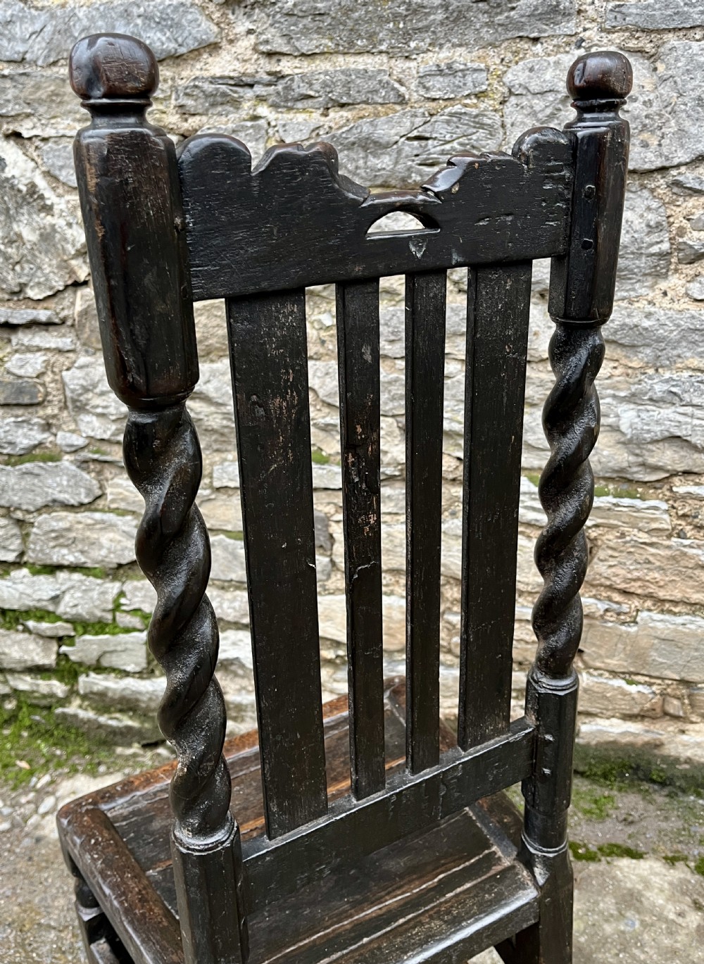 A dark 17th-century carved oak backstool with barley-twist uprights, a carved cresting rail, vertical back splats, a solid seat, and turned legs. View IX