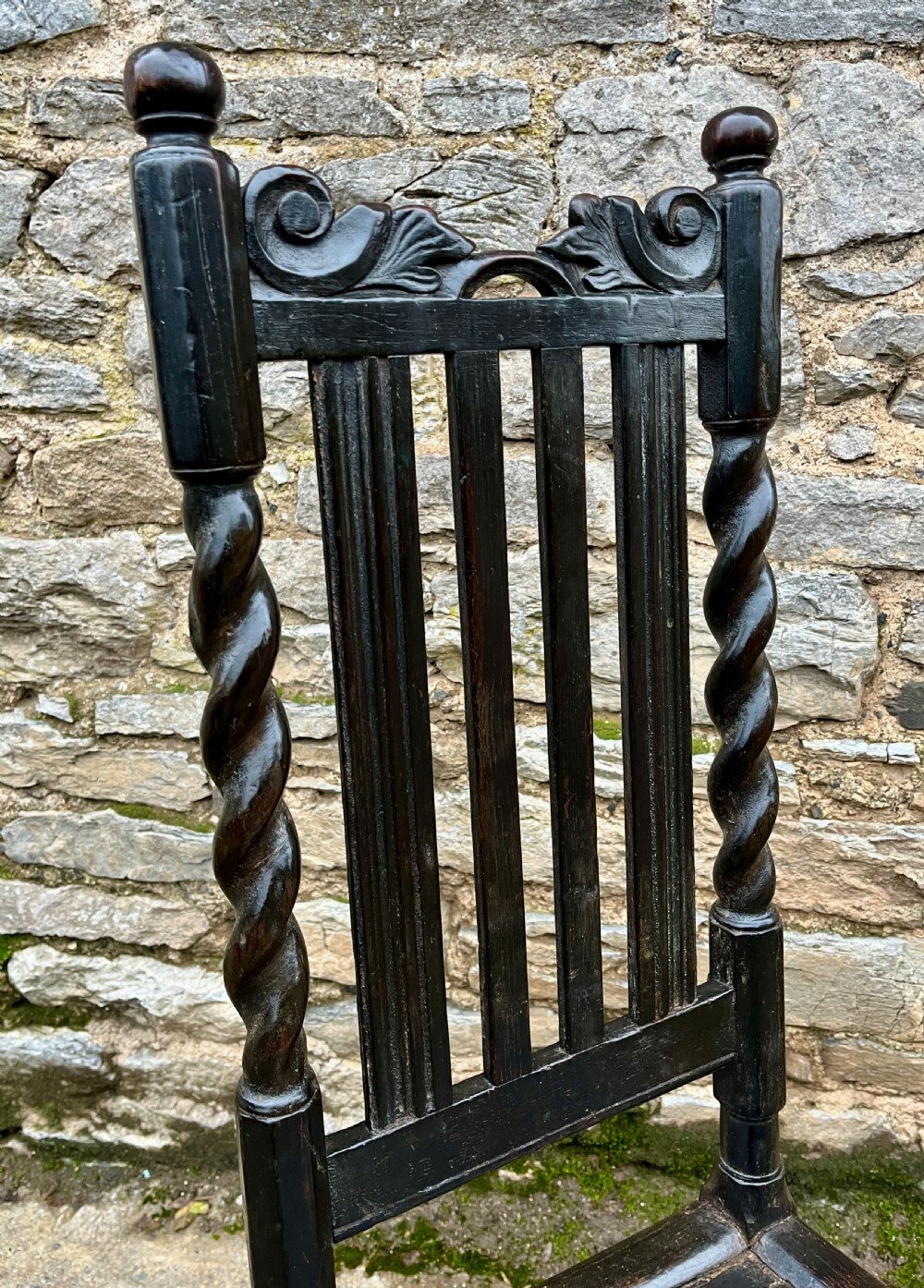A dark 17th-century carved oak backstool with barley-twist uprights, a carved cresting rail, vertical back splats, a solid seat, and turned legs. View XVII