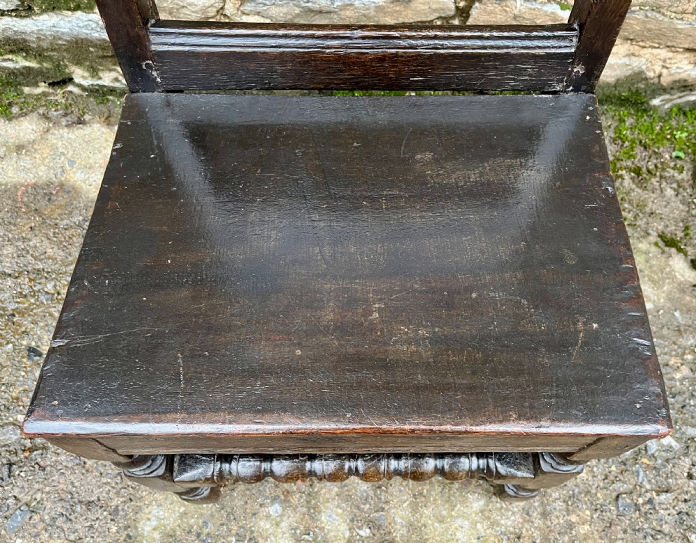A dark oak 17th-century backstool featuring a carved crested top rail, solid wooden seat, turned front legs, and a stretchered base. View III