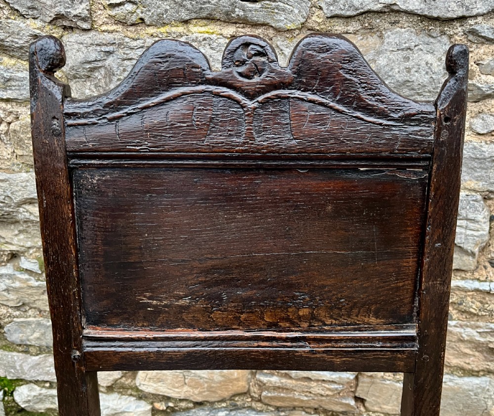 A dark oak 17th-century backstool featuring a carved crested top rail, solid wooden seat, turned front legs, and a stretchered base. View VII