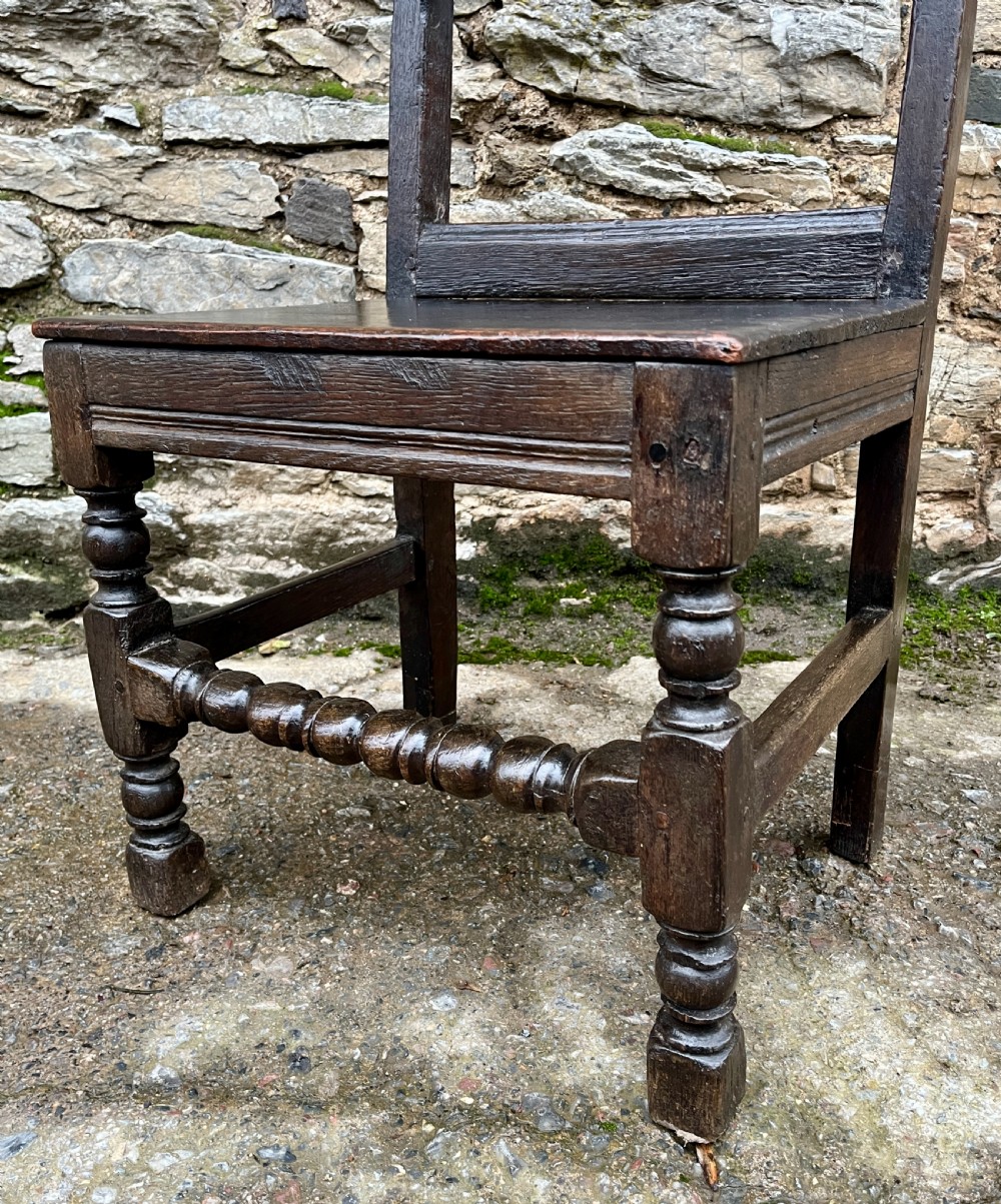 A dark oak 17th-century backstool featuring a carved crested top rail, solid wooden seat, turned front legs, and a stretchered base. View IX